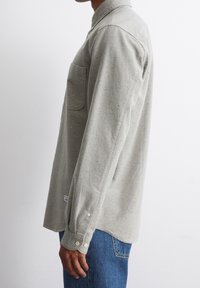 Seitenansicht einer Person, die ein hellgraues Langarmhemd mit Brusttasche und blaue Jeans vor einem schlichten weißen Hintergrund trägt.