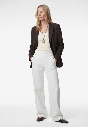 Mujer con blazer negro, camiseta beige, pantalones blancos de tiro alto con botones dorados y zapatos planos marrón oscuro, de pie con las manos en los bolsillos.