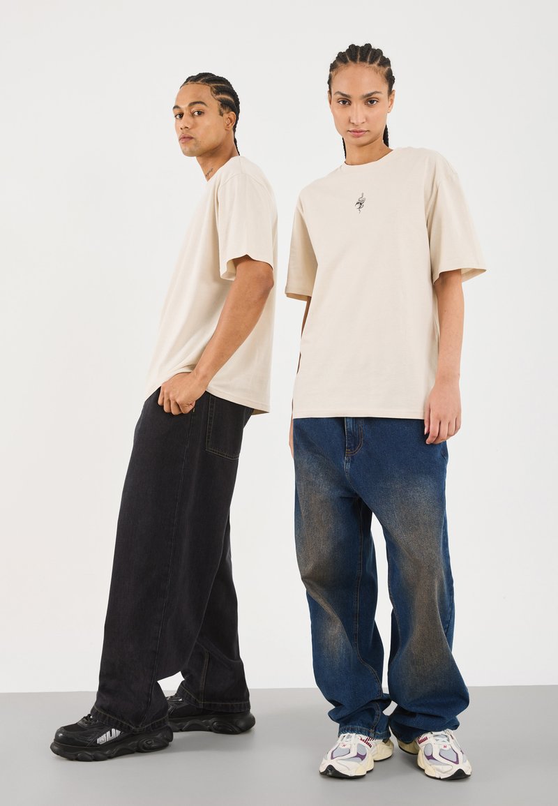 Beige T-Shirt mit kurzen Ärmeln an einem Model, kombiniert mit lockeren schwarzen und blauen Jeans. Sneaker mit mehrfarbigen Akzenten runden den Look ab.