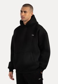 Zwarte hoodie van zacht stof met een buidelzak aan de voorkant, geribbelde manchetten en een capuchon met trekkoord. Voorzien van een klein logo op het borstgebied.