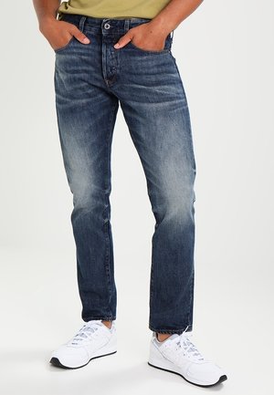 Person trägt verblasste blaue Jeans mit den Händen in den Taschen und weiße Sneakers, steht vor einem schlichten hellen Hintergrund.