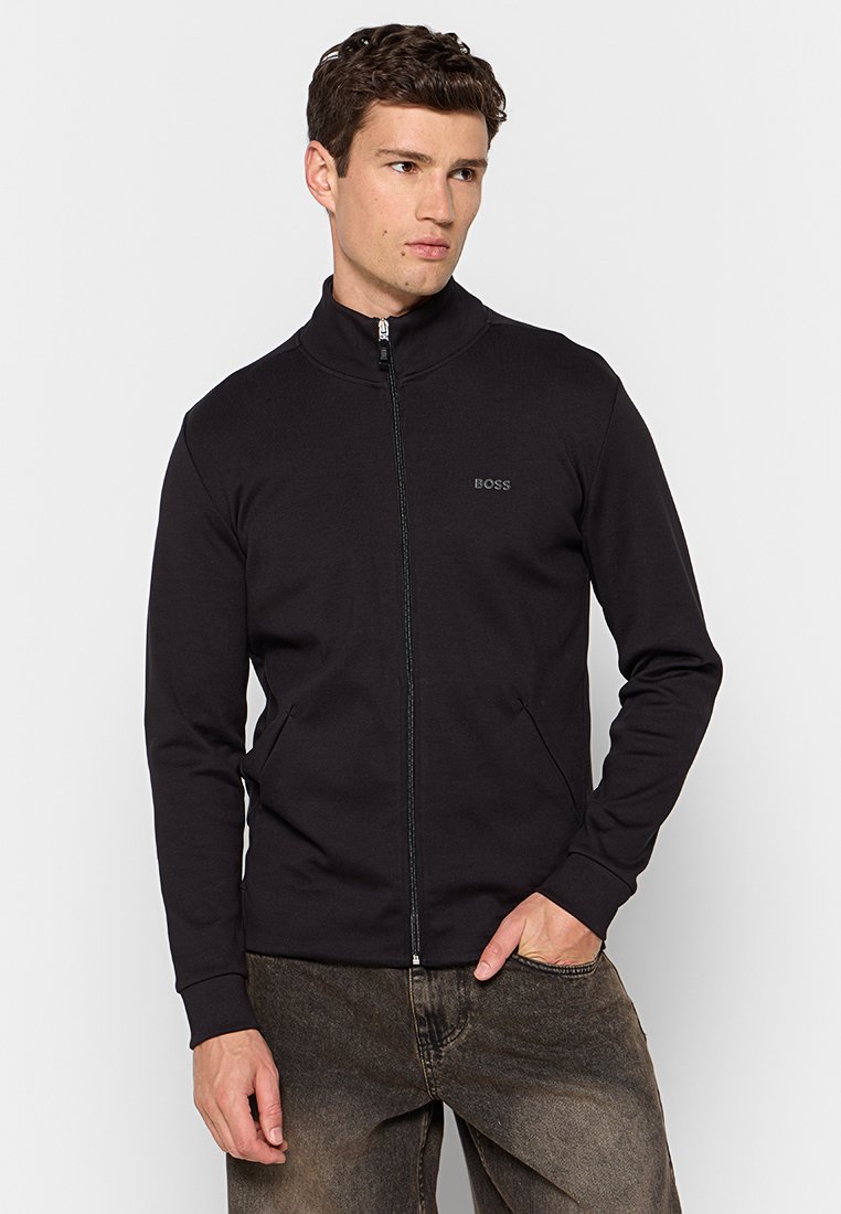 Giacca nera con zip e colletto alto, realizzata in materiale morbido, presenta tasche laterali e un logo discreto sul petto, dal taglio su misura.