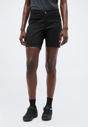 Isik, kes kannab musta keskkõrgete lühikeste pükste, musti sokke ja musti spordijalatseid, seisab ühevärvilise tausta ees.