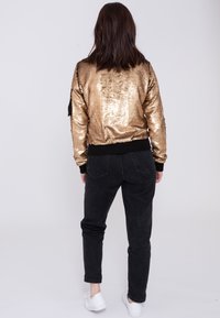 Gouden pailletten bomberjack met zwarte ribgebreide manchetten en gevel, gecombineerd met zwarte denim jeans en witte sneakers. Achteraanzicht, vrouw met middellang haar.