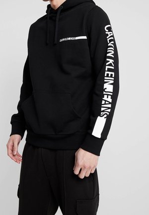 Osoba nosící černou mikinu Calvin Klein s klokáním kapsou, bílým logem na hrudi a velkým bílým nápisem na levém rukávu.