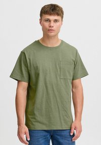 Camiseta de manga corta en verde oliva hecha de algodón texturizado. Presenta un cuello redondo y un bolsillo cuadrado en el lado izquierdo del pecho.