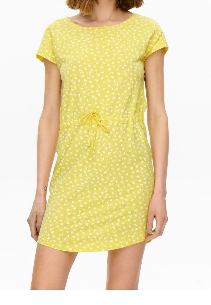 Femme portant une robe jaune à manches courtes avec un petit motif floral blanc et une taille à cordon, debout devant un fond uni.