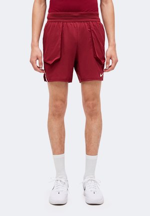 Mand iført mørkerøde sportsshorts med store forlommer, hvide crew-strømper og hvide Nike-sneakers, der står mod en enkel baggrund.