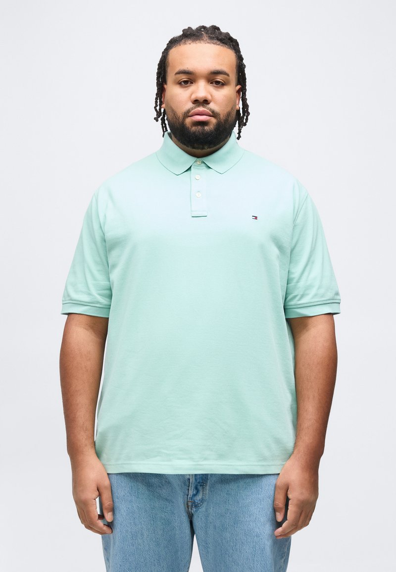 Άνδρας με πλεξούδες στα μαλλιά και γενειάδα, ντυμένος με φανελάκι τύπου πόλο ανοιχτού πράσινου χρώματος και μπλε τζιν παντελόνι, στέκεται μπροστά σε λευκό απλό φόντο.