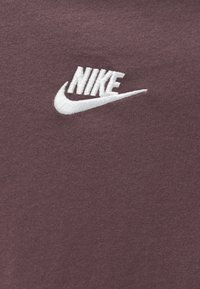 Λευκό κεντημένο λογότυπο Nike σε υφή σκούρου μωβ φόντου, που δείχνει την επωνυμία και το σύμβολο Swoosh.