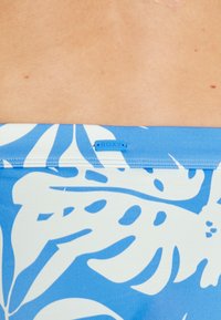 Tecido estampado com flores azuis e brancas, com uma textura suave, apresentando um rótulo da Roxy cosido na costura na parte superior.