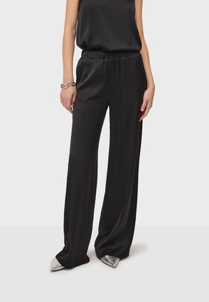 Frau trägt lockere schwarze Satin-Hose und ärmelloses Oberteil mit silbernen spitzen Schuhen und Kettenarmband, steht vor einfarbigem Hintergrund.