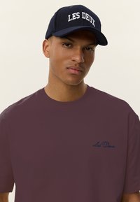 Tamsiai raudonas trumpomis rankovėmis marškinėlis su mėlynu siuvinėtu „Les Deux“ logotipu ant krūtinės, derinamas su tamsiai mėlyna beisbolo kepure su baltu užrašu.