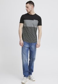 Camiseta negra y gris con bloques de color horizontales y detalle de bolsillo, combinada con jeans azul claro y zapatillas blancas. Diseño simple.