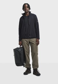 Sort fleecejakke med høj krave, sort t-shirt under, olivengrøn cargobukser og sorte vandrestøvler. Holder en sort vandtæt rygsæk.