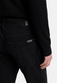 Jeans in denim nero con vestibilità slim, dotati di tasche posteriori, etichetta con logo e dettagli di cucitura discreti. Texture liscia, design minimal.