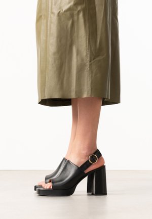 Vrouw die zwarte leren open-toe sandalen met hak en enkelbandjes draagt en een olijfgroene midirok tegen een effen achtergrond.