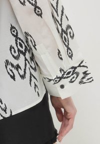 Blusa bianca con motivo astratto nero; presenta polsini abbottonati e una texture liscia; le maniche sono parzialmente visibili accanto a un indumento nero.