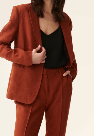 Femme portant un blazer sur-mesure couleur rouille et un pantalon assorti avec un haut noir en V, posant avec une main dans la poche et l'autre tenant le blazer.