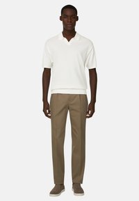 Homme portant un polo blanc à manches courtes, un pantalon beige ajusté et des chaussures marron sans lacets, debout devant un fond uni.