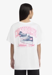 Balta per didelį T-shirts su mėlynomis sportinėmis batelėmis, ant nugaros ryškiai raudonas užrašas "CONVERSE" ir "Sporto skyrius".