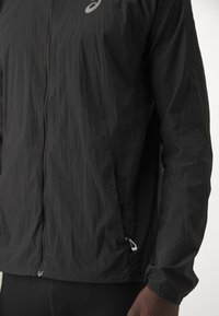 Mees, kes kannab musta kerget jope ees luku ja Asicsi logoga rinnal, kujutatud torso ja randme vahelt.