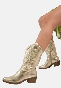 Botas de vaquero de cuero dorado con diseño texturizado, punta puntiaguda y tacón bajo. Cuenta con un patrón embosado y cierre de cremallera.