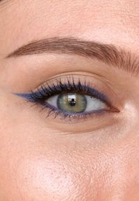 Nærbilde av et grønt øye med blå eyeliner. Huden er jevn med en lys tone, og øyevippene er lange og mørke. Fint formet bryn er synlig.