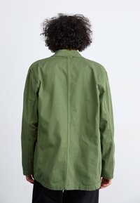 Grønn bomullsjakke med rett snitt, lange ermer og midt bak søm. Enkel design, ingen synlige deler eller mønstre.