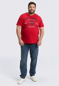 Rode katoenen T-shirt met zwarte tekst, uitgevoerd met een ronde hals en een ontspannen pasvorm. Gecombineerd met blauwe spijkerbroek en witte sneakers.