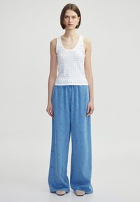 Weißes ärmelloses Tanktop mit kleinen Pünktchen, kombiniert mit weit geschnittenen hellblauen Jeanshosen mit floralen Stickereien.