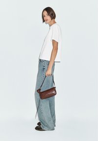 Maglietta bianca a maniche corte, jeans a gamba larga blu chiaro, e borsa in pelle marrone con tracolla, indossando scarpe nere su uno sfondo semplice.