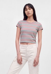 Giovane donna con lunghi capelli scuri che indossa un crop top a righe rosa e verde e pantaloni cargo bianchi, in piedi contro uno sfondo semplice.