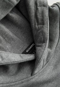 Gros plan sur un tissu gris montrant un col de vêtement avec une étiquette visible de la marque AllSaints en noir à l'intérieur.