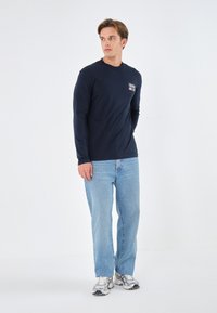 Marineblaue Langarmshirts mit kleinem Logo auf der Brust, kombiniert mit hellblauen Jeans und silbernen Sneakers, stehend vor einem schlichten Hintergrund.