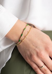 Złote bransoletki z zielonymi akcentami; jedna ma wydłużone sekcje, podczas gdy druga ma małe koralikami na łańcuszku.