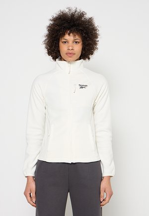 Frau mit lockigem Haar, die eine weiße Reebok Fleecejacke mit Reißverschluss und dunkelgraue Hose trägt, steht vor einem einfachen hellen Hintergrund.