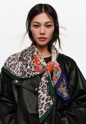 Mujer con cabello oscuro que lleva una chaqueta de cuero negra y una bufanda de seda de múltiples patrones atada al cuello, sobre un fondo blanco liso.