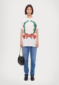 Bílý krátký rukáv tričko s grafikou zeleného vavřínového věnce, červenou stužkou a textem "Moschino". V kombinaci s modrými džíny a černou kabelkou.