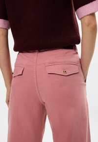 Ροζ βαμβακερές παντελόνες με πίσω τσέπες και χρυσά κουμπιά. Φοριούνται με ένα βουργουνδί και ροζ πουλόβερ.