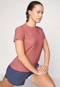 Camisa de desporto de manga curta em rosa suave, com textura lisa, apresentando um logótipo no peito. Combinada com calções azul escuro, revelando um aspeto desportivo.