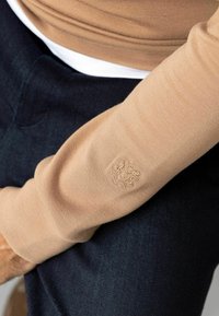 Maglione beige a maniche lunghe con texture a coste, dotato di un piccolo logo ricamato sulla manica. Abbinato a jeans denim blu scuro.