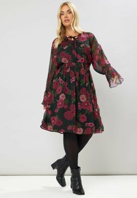 Abito floreale con sfondo nero, caratterizzato da rose rosa e rosse. Tessuto fluido e trasparente con maniche lunghe a campana. Stivaletti neri alla caviglia completano il look.