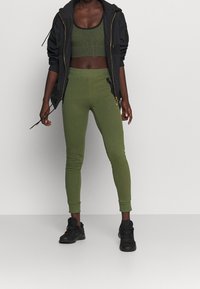 Sort zip-up jakke, grøn ribbet crop top, grøn tætsiddende leggings og sorte sneakers. Leggings har ankelmanchetter; jakken har hætte.