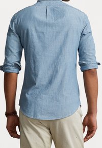 Camicia di jeans blu chiaro con maniche lunghe arrotolate, caratterizzata da una piega sul retro e un orlo curvo. Indossata con pantaloni chiari e strappati.