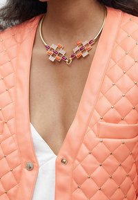 Gul chokerhalsband med fyrkantiga mönster i röda, lila och orange stenar. Bärs med en vadderad koralljacka med nitar.