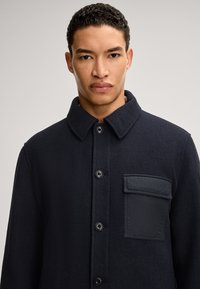 Veste en mélange de laine navy avec col, boutons à l'avant et une poche plaquée dotée d'un détail en tissu noir lisse. Surface texturée sur l'ensemble.