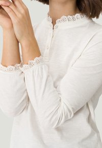 Biała bluzka z długim rękawem, ozdobiona koronczanym wykończeniem w falbanki przy kołnierzyku i mankietach. Miękka, lekka tkanina o luźnym kroju. Dwa guziki przy szyi.