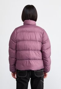 Lila pufferjacka med vertikal quiltad design, hög krage och elastiska cuffs. Mjuk, slät textur med en något glänsande yta.