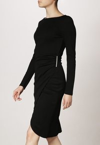 Černé šaty s dlouhým rukávem, s přiléhavým střihem, bočními řasením a ozdobným detailem v pase. Hladká textura a délka ke kolenům.